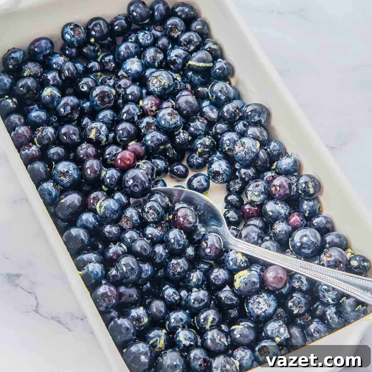 Classic Blueberry Cobbler Recipe with Cinnamon Crumble 5 A rectangular white dish filled with fresh, dark blueberries, with a silver spoon resting inside. The dish is placed on a light marble surface.