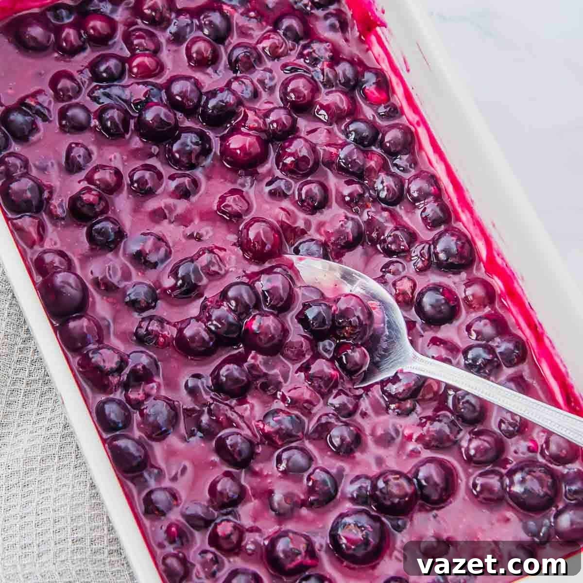 Classic Blueberry Cobbler Recipe with Cinnamon Crumble 6 A rectangular white dish filled with glossy baked blueberries, with a silver spoon resting inside, surrounded by a textured gray cloth on a white surface.