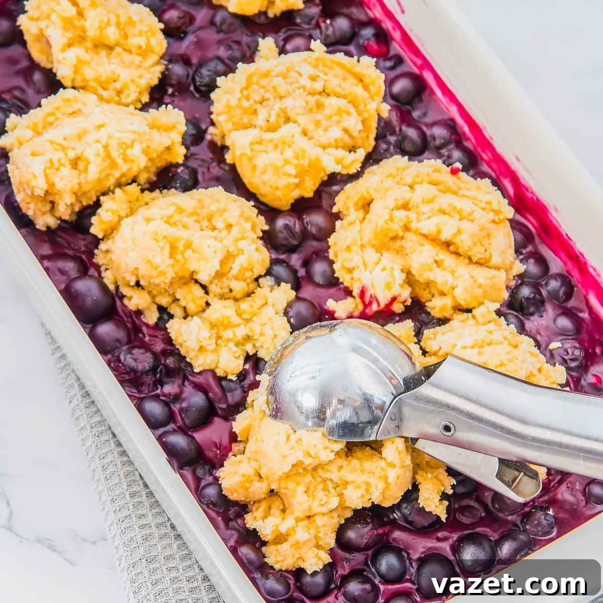 Classic Blueberry Cobbler Recipe with Cinnamon Crumble 10 A baking dish filled with blueberries topped with scoops of yellow biscuit dough, placed with a metal ice cream scoop, ready to be baked.