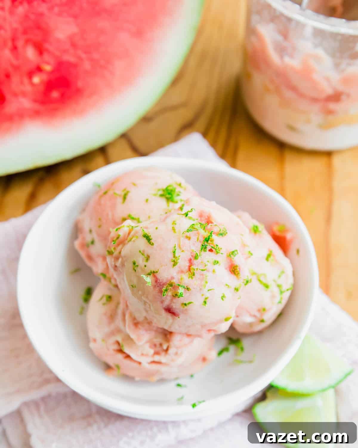 Creamy Watermelon Ice Cream Recipe 2 Three scoops of watermelon ice cream garnished with lime zest in a white bowl, with lime slices and a jar of ice cream nearby on a wooden surface. A watermelon is visible in the background.