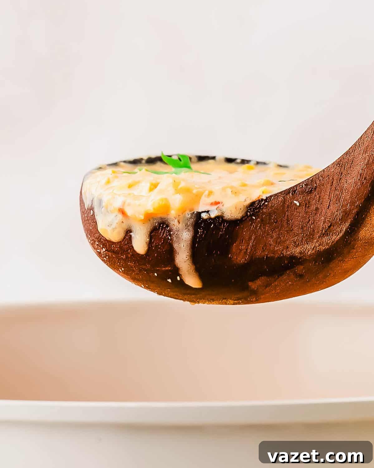 A wooden ladle holds creamy soup with corn and herbs, dripping slightly over the edge, above a white bowl.