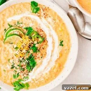 Creamy corn soup garnished with grilled corn, lime slices, jalapeño, fresh cilantro, crumbled cheese, and a swirl of cream, served in a white bowl with a spoon nearby.