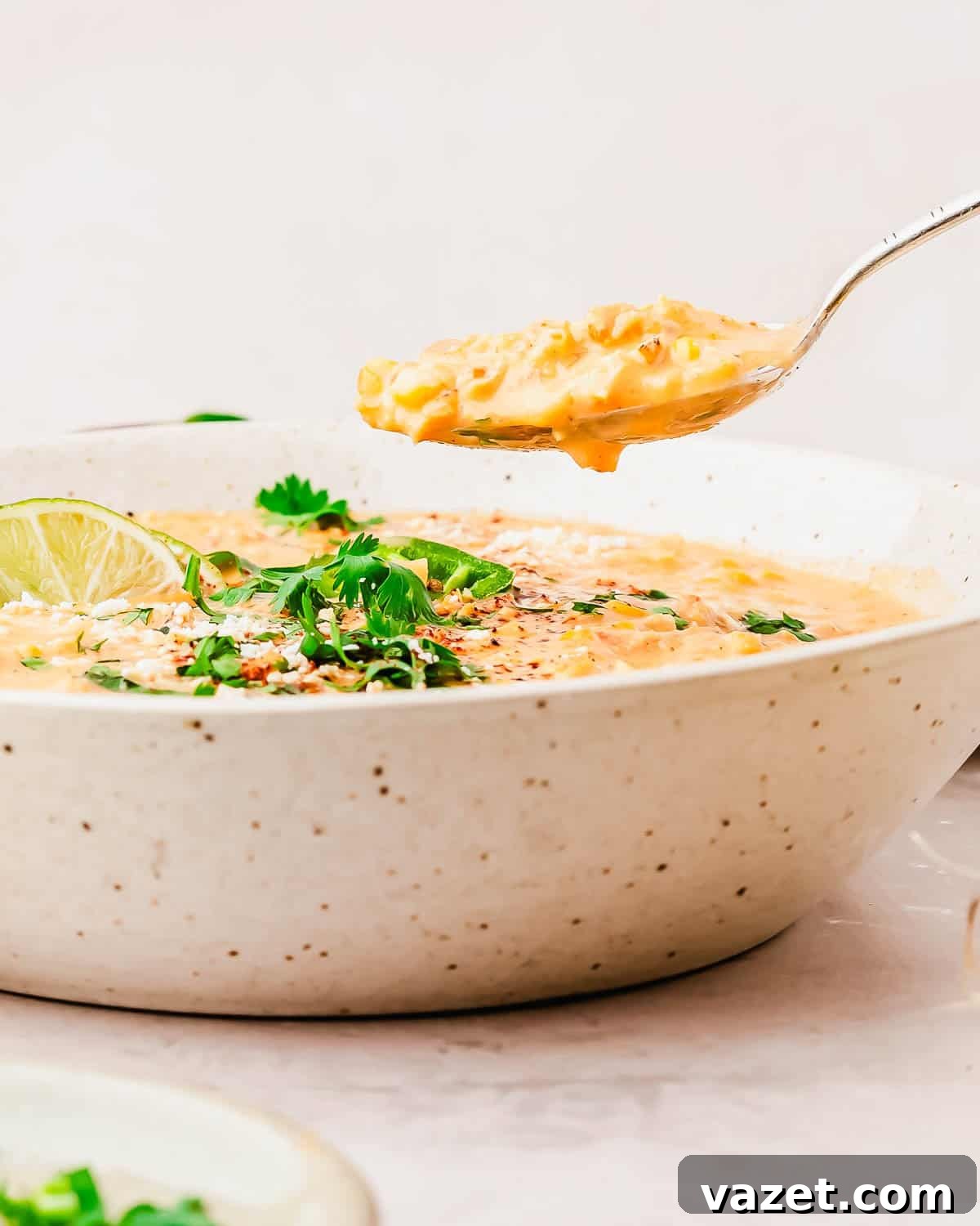 A close-up of a bowl of creamy street corn soup garnished with cilantro, lime, and sesame seeds, with a spoon lifting a bite from the bowl.
