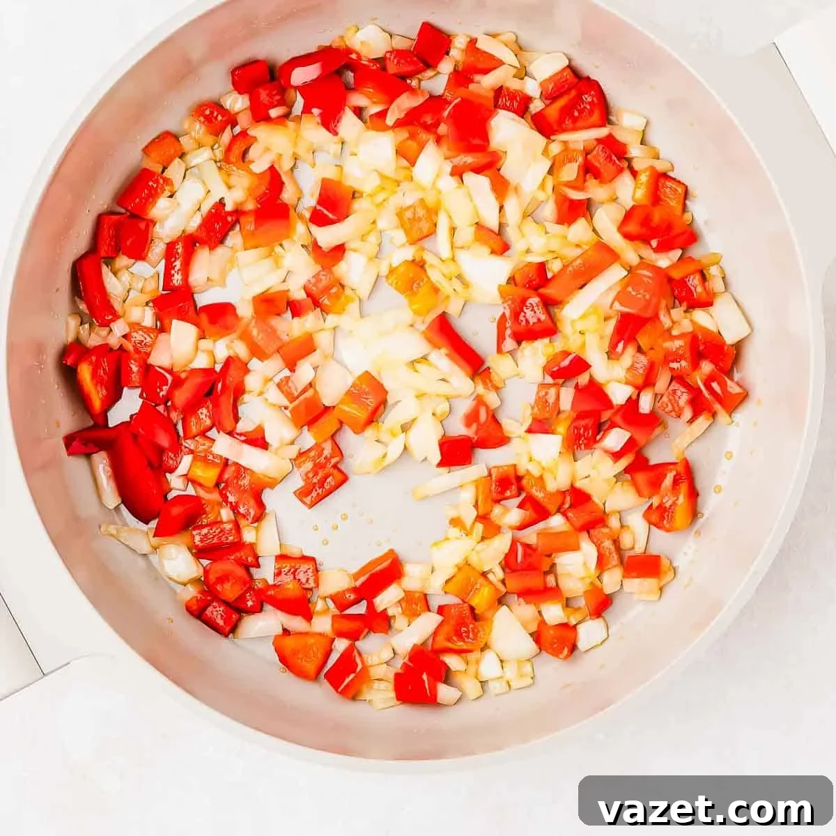 Diced red bell peppers and onions are being sautéed in a white skillet.