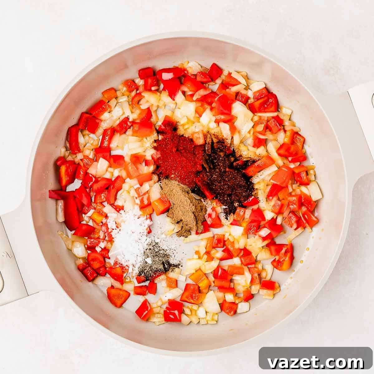 A white pot with chopped red bell peppers, onions, and piles of spices including black pepper, salt, paprika, and cumin, arranged before mixing.