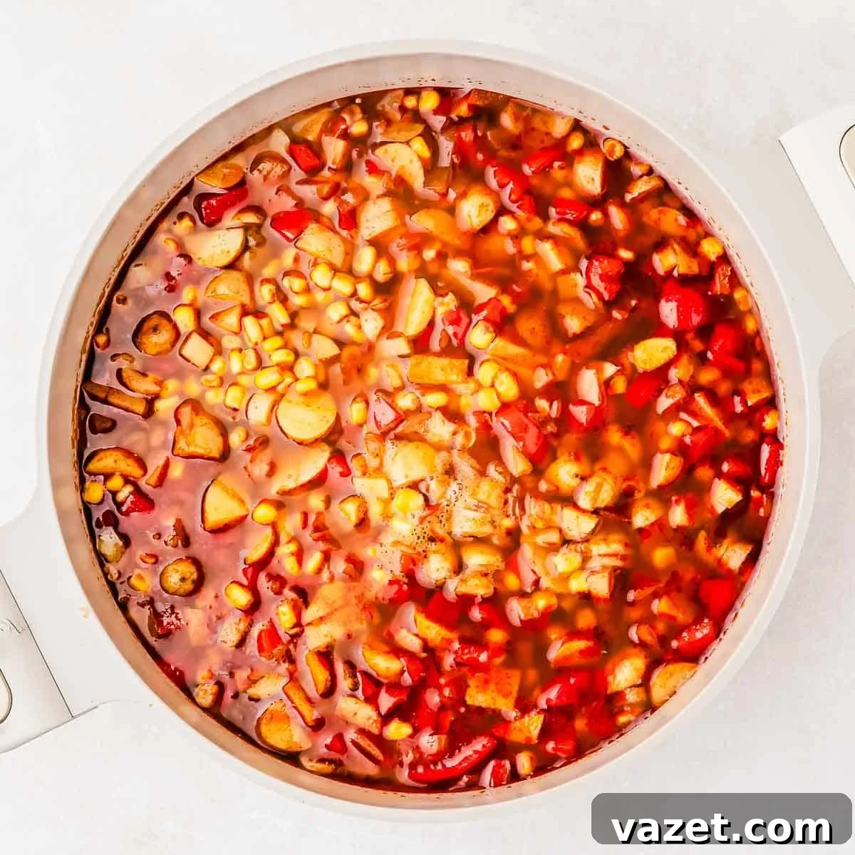 A pot filled with a colorful soup, featuring visible corn, red bell peppers, potatoes, and a rich red broth.