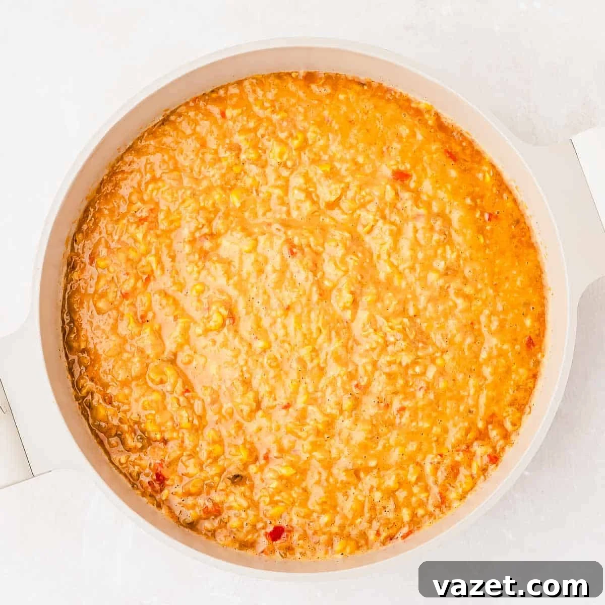 A round white pot filled with creamy orange corn soup featuring visible small bits of vegetables, sits on a light surface.