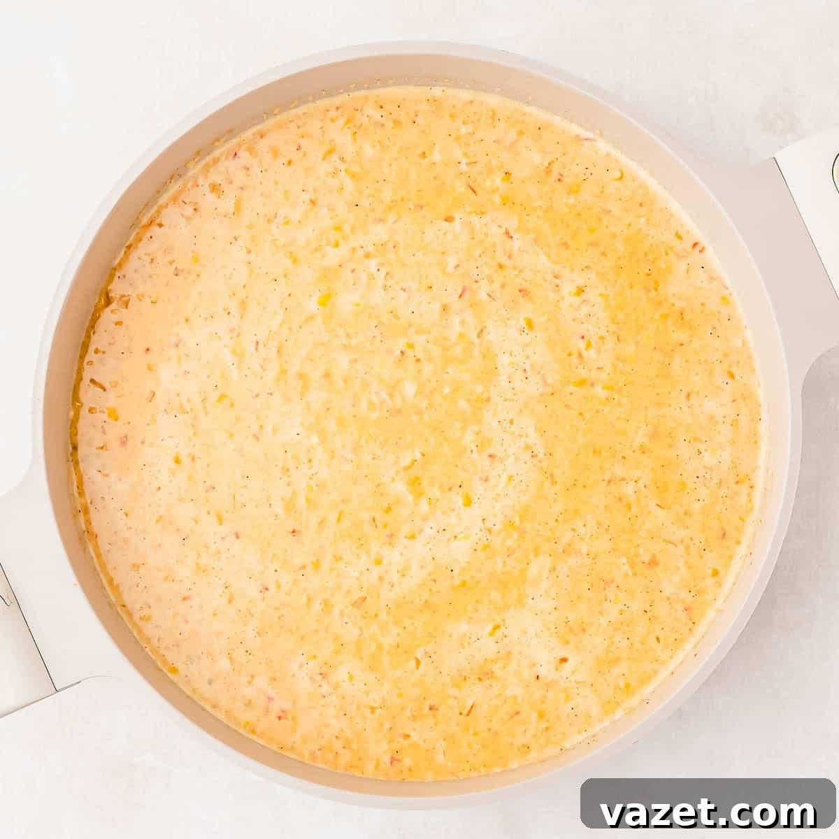 A creamy, yellow corn soup simmering in a light-colored pot, viewed from above on a light surface. Small bits of herbs and ingredients are visible throughout the soup.