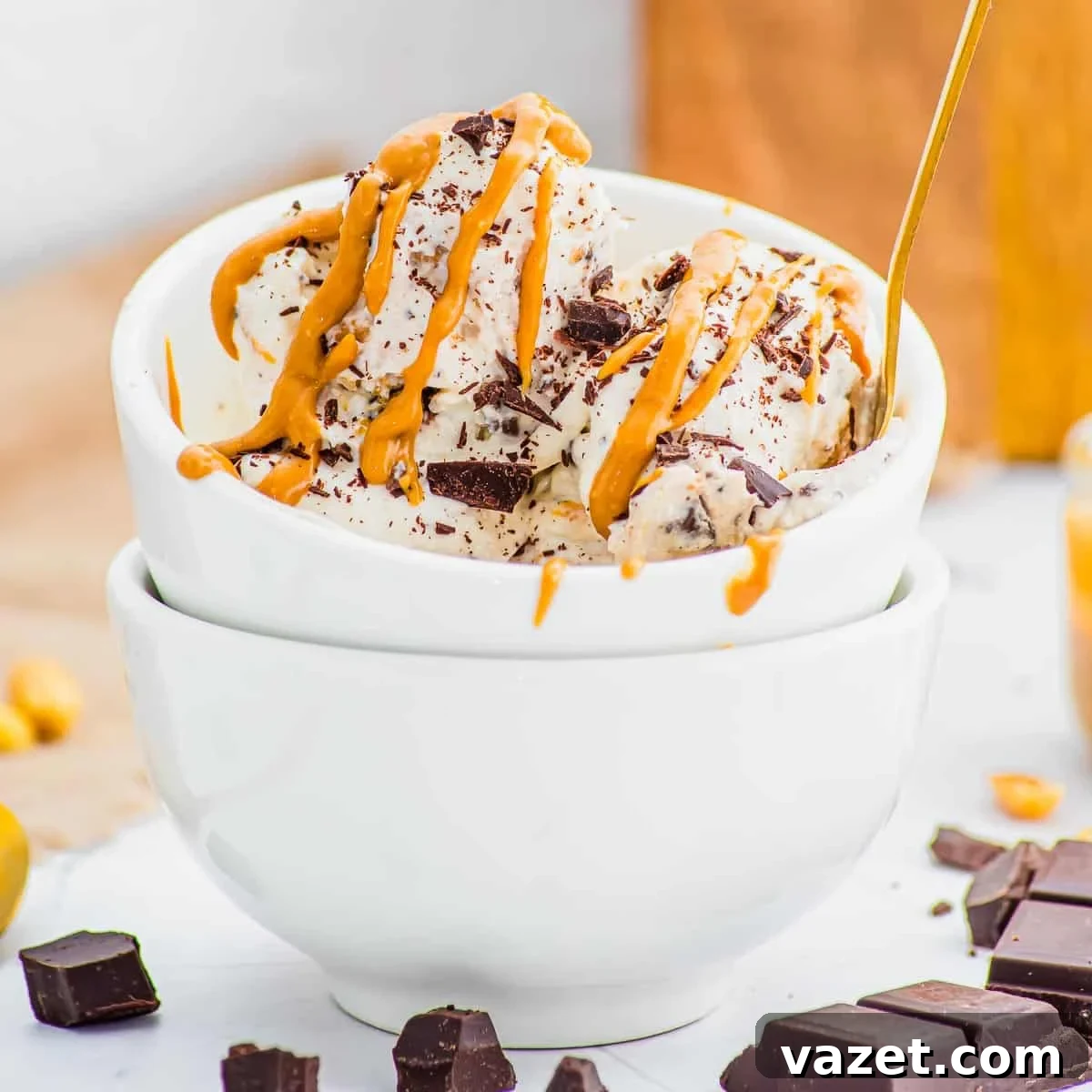 Two scoops of ice cream with chocolate chunks and peanut butter drizzle in a white bowl, stacked on another bowl, with pieces of chocolate nearby.