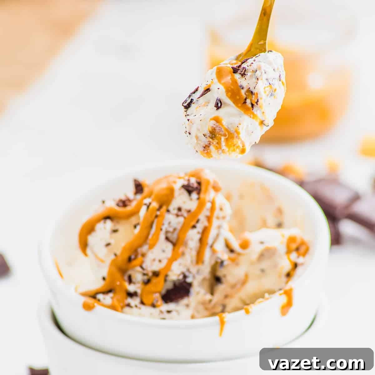 A spoonful of creamy cottage cheese ice cream with chocolate chunks and peanut butter drizzle is held above a white bowl filled with more ice cream, with extra peanut butter and chocolate pieces nearby.