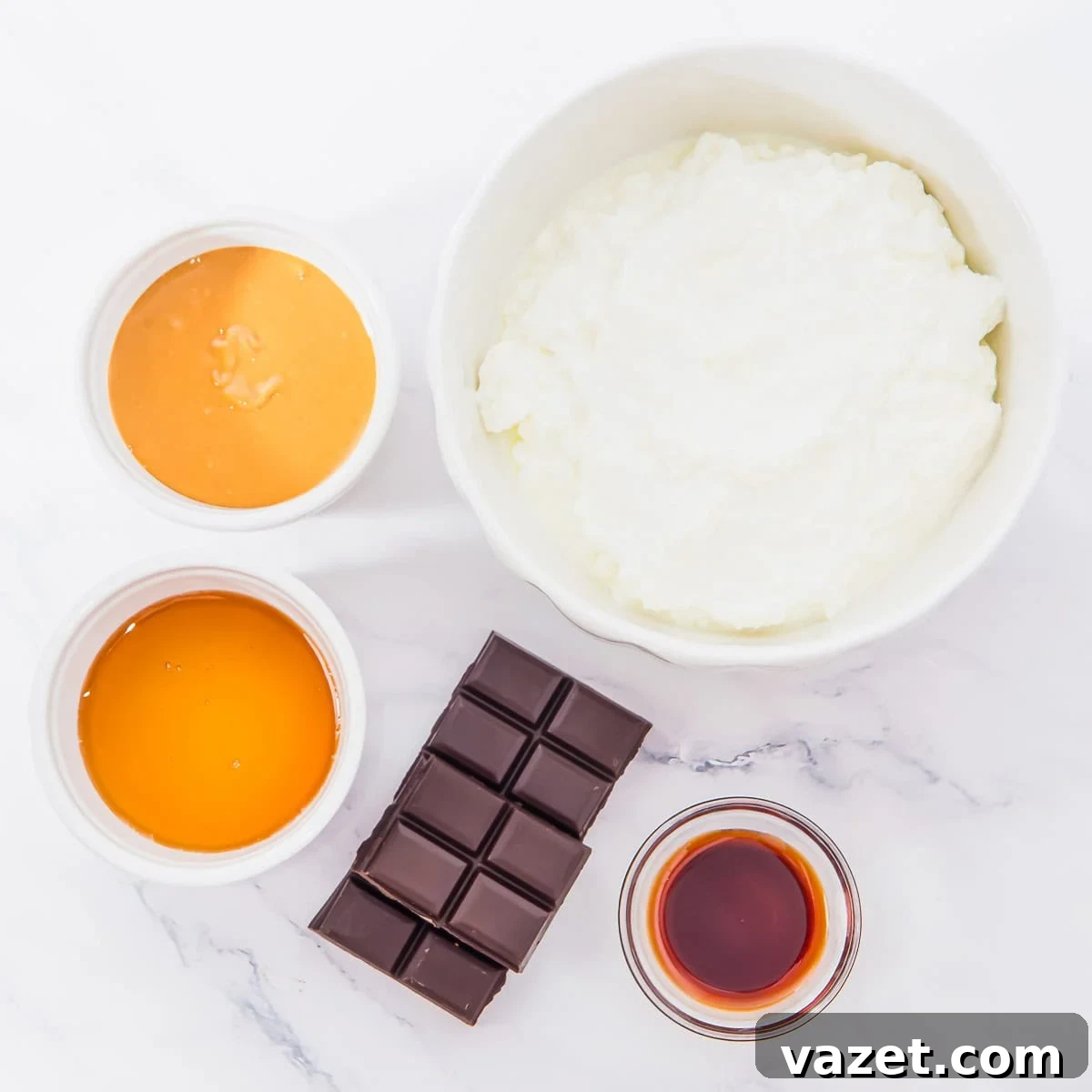 A bowl of cottage cheese, a bar of dark chocolate, small bowls of peanut butter, honey, and vanilla extract on a white marble surface.