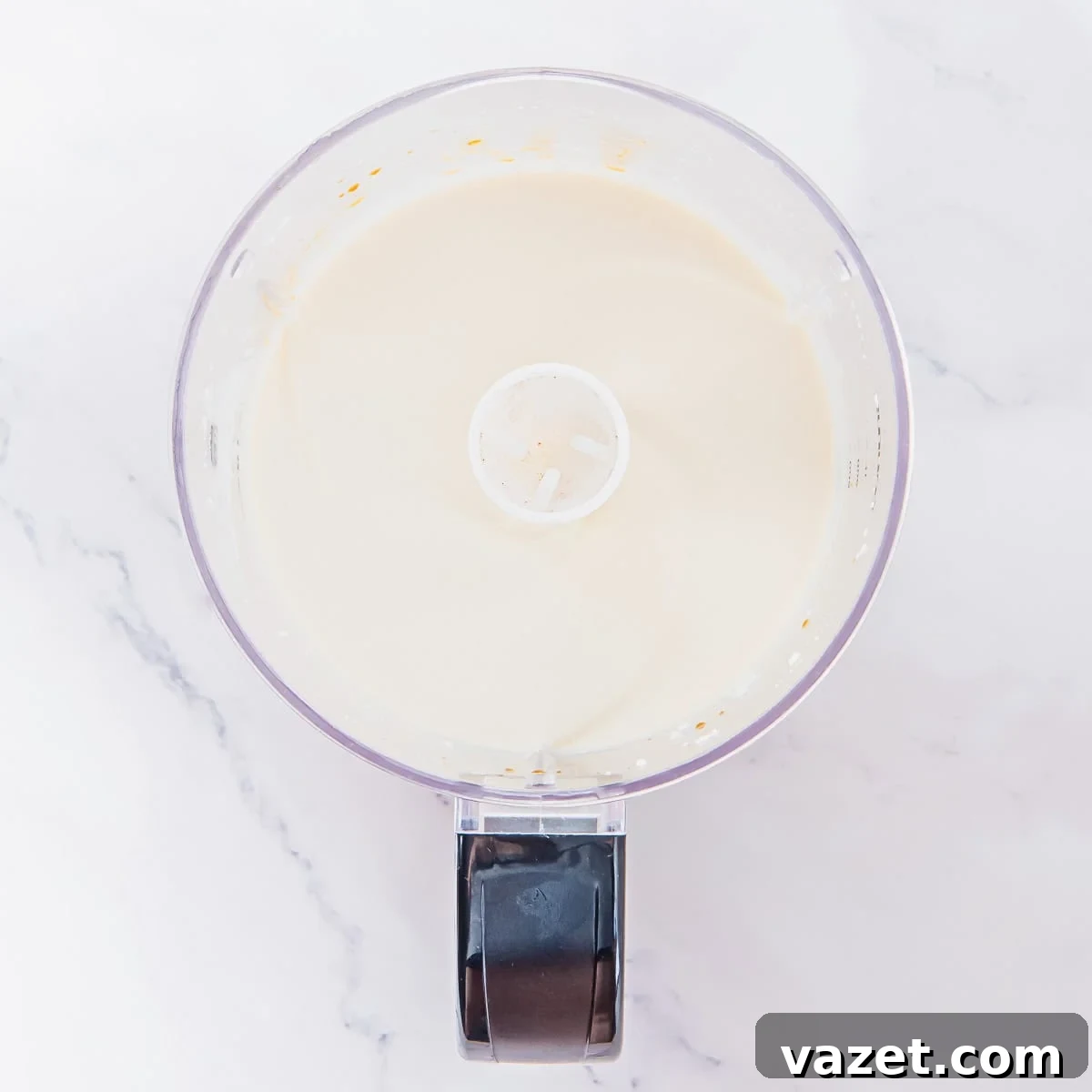 A food processor bowl filled with smooth, creamy whipped cottage cheese mixture sits on a white marble countertop, viewed from above.