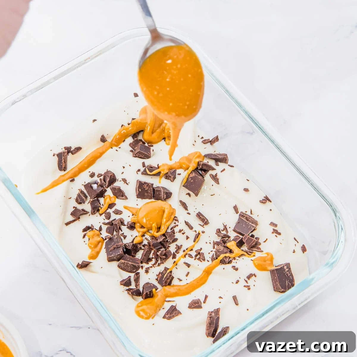 A spoon drizzles peanut butter over a glass dish filled with creamy whipped cottage cheese, topped with chocolate chunks and more peanut butter, on a white surface.