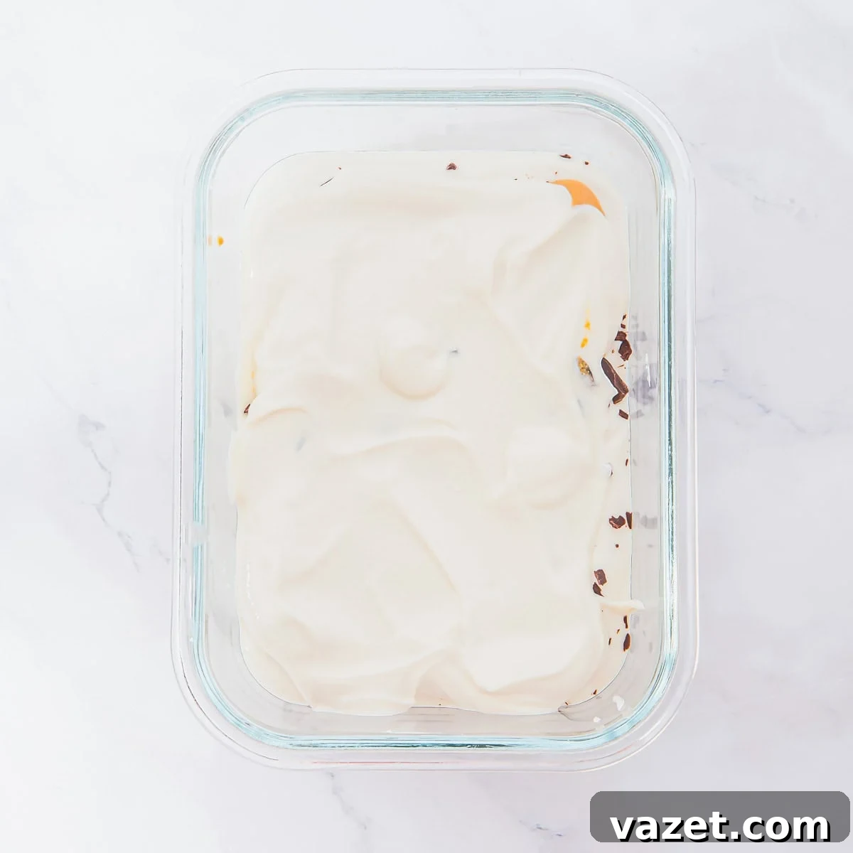 A glass container filled with a layer of creamy white cottage cheese covering some chocolate pieces, sitting on a white marble surface.