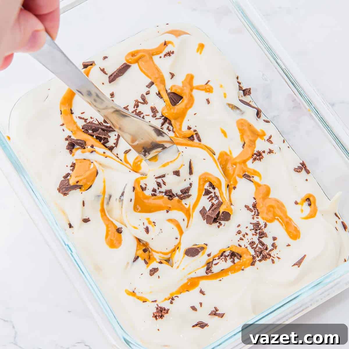 A hand uses a spoon to swirl peanut butter and chocolate shavings into a creamy ice cream mixture in a glass dish on a light surface.