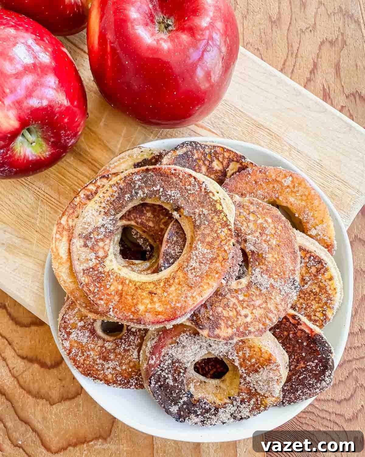 A plate of sugar-coated apple ring fritters sits on a wooden board, with two whole red apples nearby.