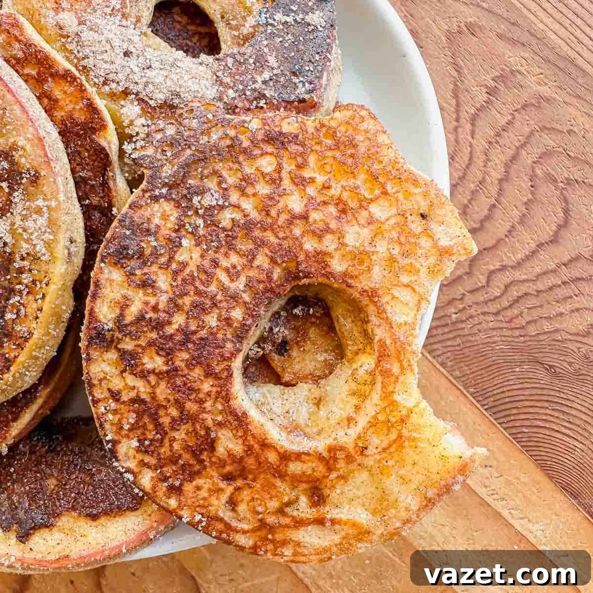 A close-up of apple slices coated in batter, fried to a golden brown, and dusted with sugar, served on a white plate atop a wooden surface. One slice has a bite taken out.