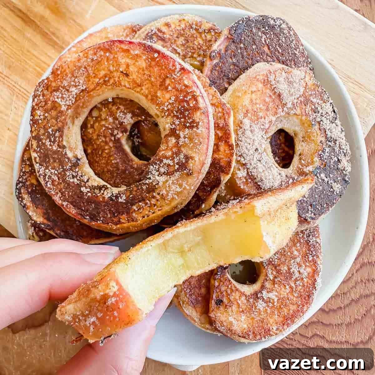 A hand holds a fried apple ring coated with cinnamon sugar above a plate stacked with more apple rings, all with holes in the center and golden brown edges.