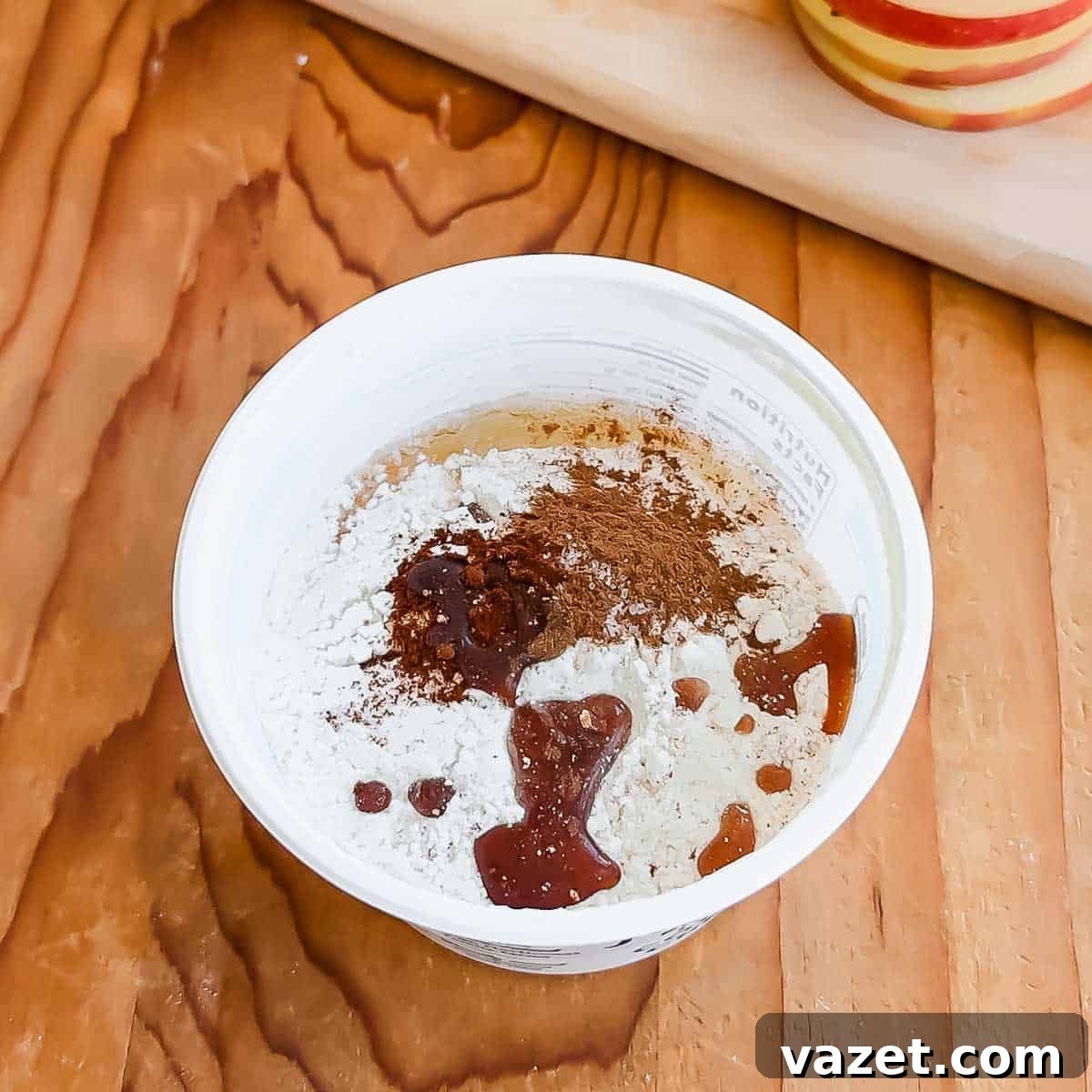 A white container filled with flour, cinnamon, and a drizzle of vanilla extract sits on a wooden surface, with a blurred cutting board and sliced apples in the background.