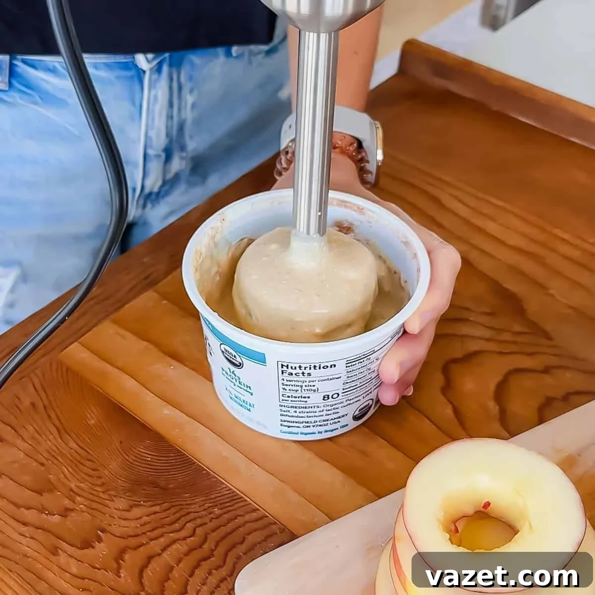 A person uses an immersion blender to blend a batter mixture in a cottage cheese container. Sliced apples with cores removed are on a cutting board nearby.
