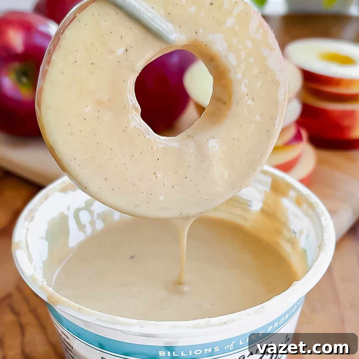 A round apple slice with a hole in the middle is being dipped into creamy batter, with apples and cottage cheese container in the background.