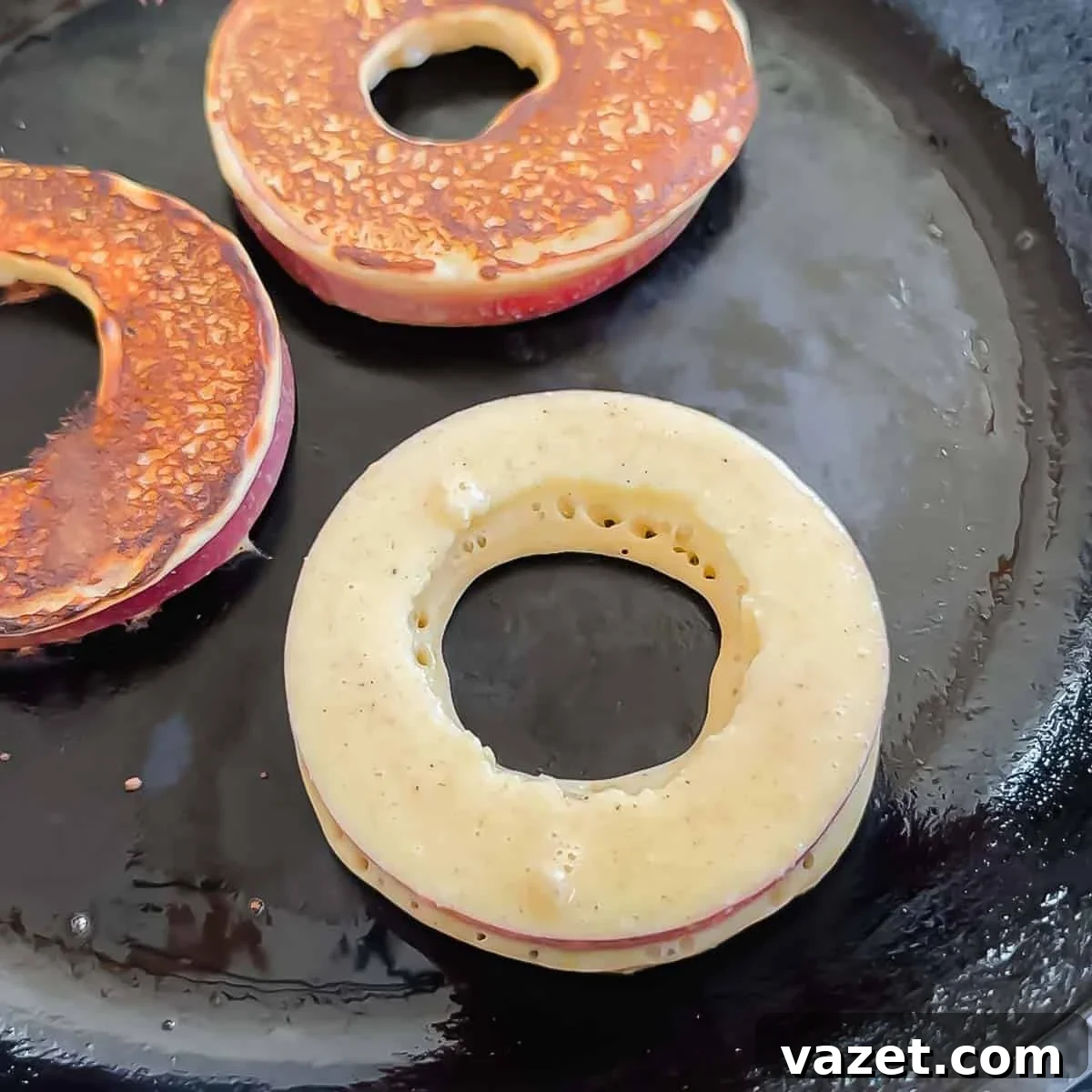 Three round apple slices dipped in pancake batter are cooking on a black skillet. Two slices are golden brown and one is still raw with visible bubbles forming in the batter.