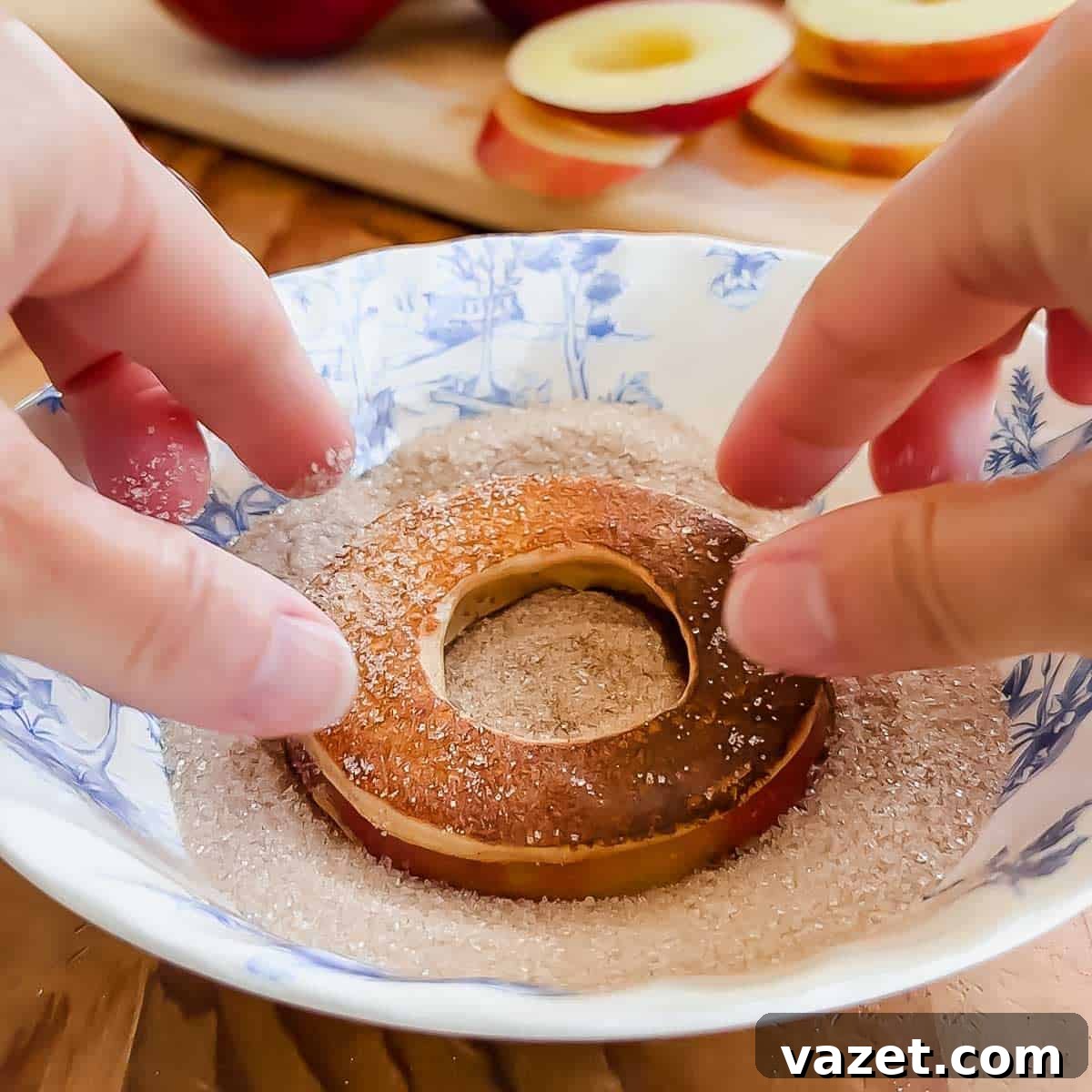 Hands dipping a round apple slice with a hole in the center into a bowl of cinnamon sugar. Sliced apples are visible on a cutting board in the background.
