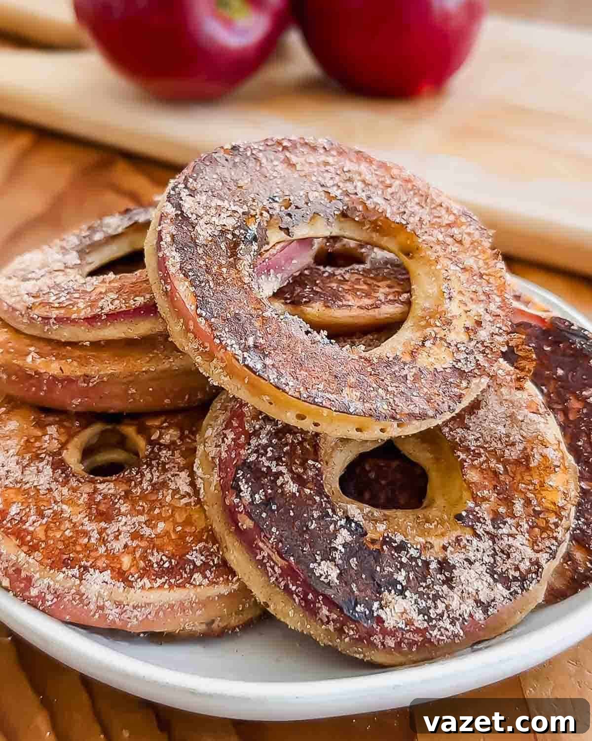 A plate of baked apple rings coated with cinnamon sugar, with red apples and a wooden cutting board in the background.